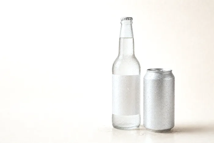 Condensation-covered blank glass bottle with a clear unprinted film label beside a sweating blank aluminum can on warm paper.