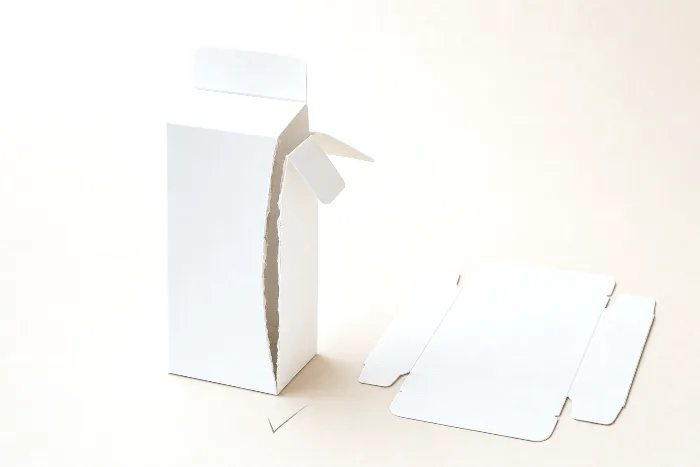 Blank white folding carton with a split, cracked crease and sprung glue flap next to a flat die-cut carton blank.