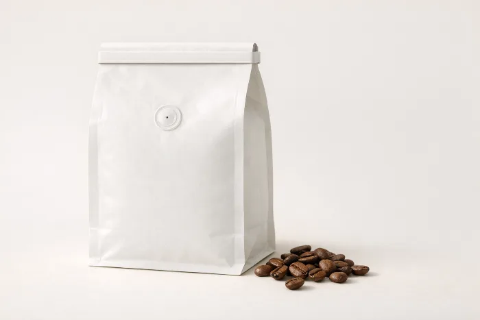 A blank kraft coffee bag with a one-way degassing valve and a fold-down tin-tie top, a few roasted coffee beans beside it.