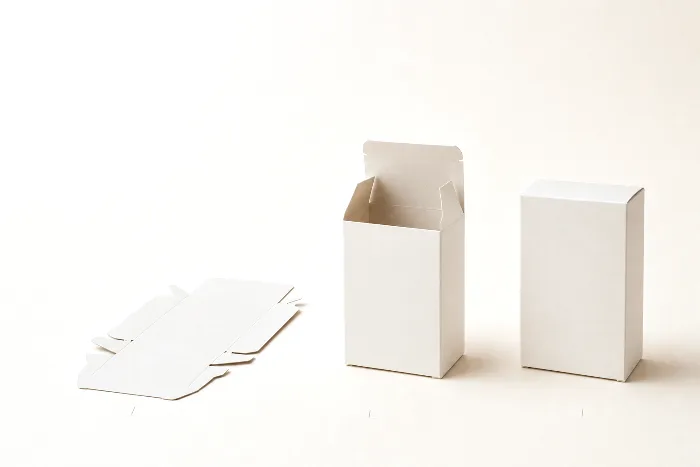 A flat carton blank, a half-folded carton, and a finished blank carton in a row on warm paper with pencil tick marks beneath.