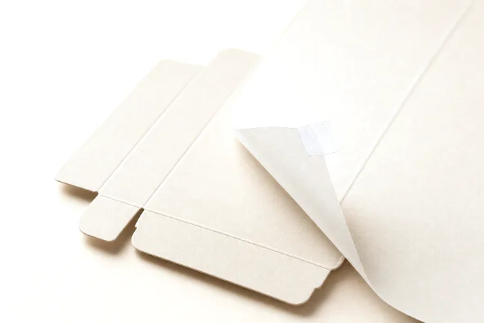Close-up of a blank die-cut carton sheet with a translucent overlay lifted at one corner, held open by a tab of clear tape.