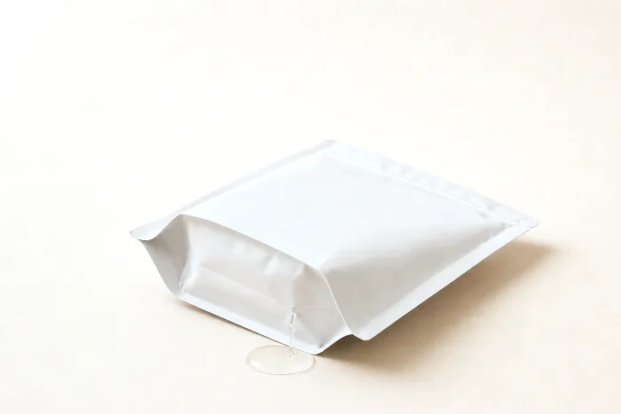 Blank white stand-up pouch on its side with water seeping from a bottom gusset corner into a small pool on the paper.
