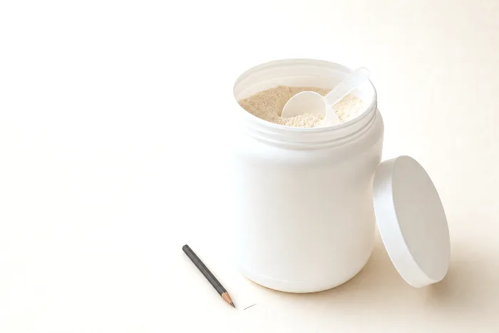 Open white powder tub two-thirds full with a scoop resting inside, visible headspace, and a pencil tick on the paper marking the fill level.