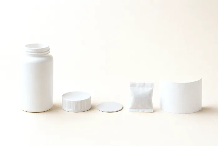 White unlabeled supplement bottle in a row with its cap, a foil liner disc, a desiccant canister, and a blank curled wrap label.