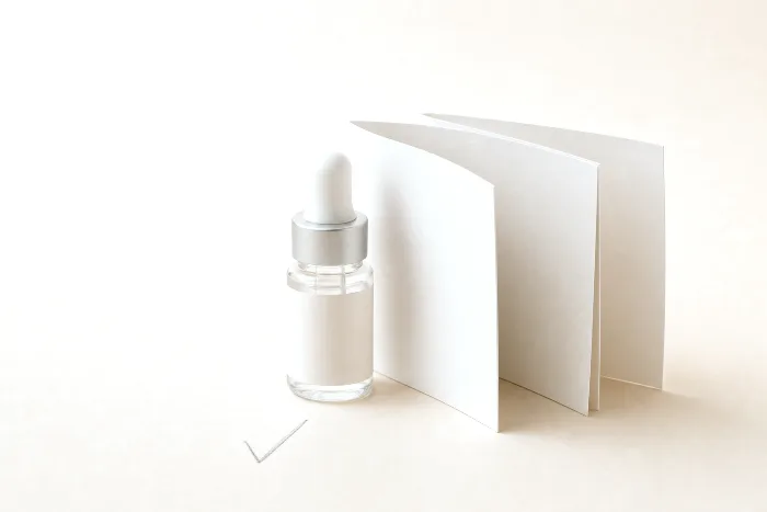 Small blank bottle next to an unprinted booklet label fanned open, with a pencil tick on warm paper marking its footprint.