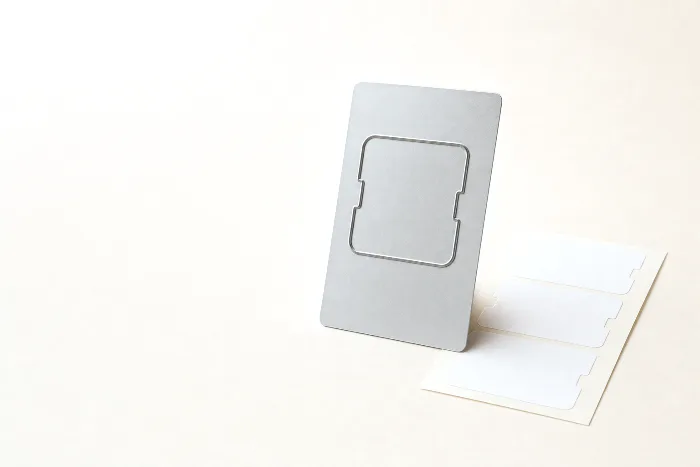 Flexible steel cutting die with a raised label-shaped blade line beside a sheet of blank rounded-corner labels, a pencil tick on the paper.