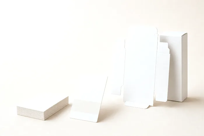 Paperboard swatch squares, an ink drawdown card, a flat die-cut blank, and a folded blank white carton in a receding row.