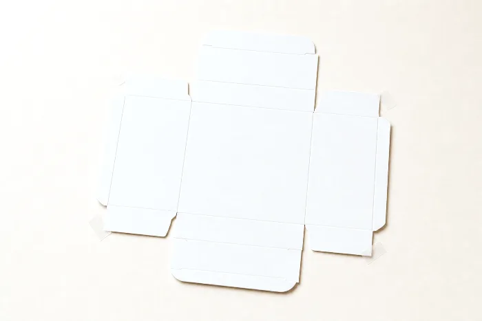 A flat unprinted die-cut carton blank on warm paper, with small tabs of translucent tape marking its corner edges.