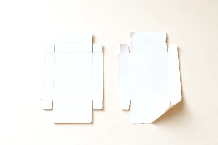 A crisp die-cut carton blank with crease lines beside a hand-cut paper copy with wavy edges and one corner curling up.